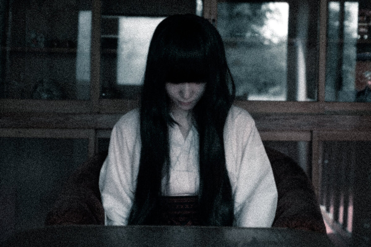 薄暗い室内で俯く長い黒髪の女性の不気味な表情